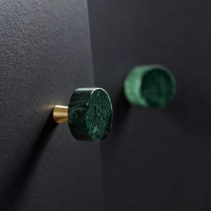 May include: Two green marble cabinet knobs with brass stems are mounted on a black surface. The knobs have a circular shape and a polished finish, showcasing the natural veining of the stone. The hardware adds a touch of luxury to any cabinetry.