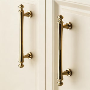 May include: Two gold-colored cabinet pulls with a vertical, elongated design. Each pull has a decorative finial at the top and bottom, and is mounted on a white cabinet door. The pulls are made of metal and have a shiny finish.