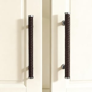 Op de afbeelding: Twee donkerbruine, geweven leren deurgrepen met zilverkleurige eindkappen zijn gemonteerd op een witte deur. De grepen zijn cilindrisch en verticaal georiënteerd, wat een vleugje elegantie toevoegt aan het ontwerp.