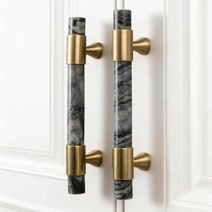 May include: Two cabinet pulls with a gray and white marble design. The pulls are cylindrical with gold-colored metal accents. The pulls are mounted on a white cabinet door.