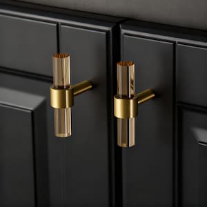 Op de afbeelding: Twee kastgrepen met een gouden afwerking en een helder, cilindrisch acryl middenstuk. De grepen zijn gemonteerd op een zwarte kastdeur. De hardware heeft een modern, elegant design.