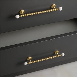 May include: Close-up of two dark gray cabinet drawers with gold-colored beaded handles. The handles have white and gray marble-like end pieces. The drawers are closed, with one slightly ajar, revealing a gray interior.