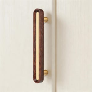 Könnte beinhalten: Ein dunkelbrauner Holzschrankgriff mit einer messingfarbenen Mitteleinlage und messingfarbenen Endkappen. Der Griff ist an einer weißen Schranktür montiert und zeigt ein modernes Design.