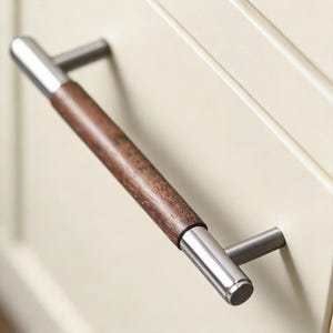 May include: A cabinet pull with a dark brown wooden center and brushed nickel ends. The handle is mounted on a cream-colored cabinet door. The pull is a modern design.