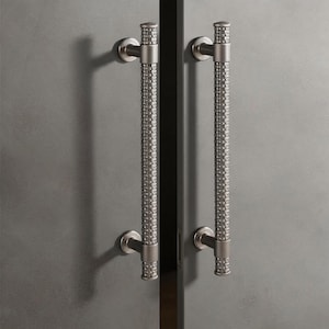 Peut inclure: Deux poignées de porte en nickel brossé avec un motif texturé et tissé. Les poignées sont cylindriques avec des embouts décoratifs et sont montées sur une surface grise. Les poignées sont symétriques et créent une esthétique moderne.