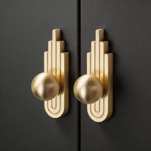 Könnte beinhalten: Zwei goldfarbene Schrankgriffe mit einem kugelförmigen Knauf und einem vertikalen Design im Art-Déco-Stil. Die Griffe sind an einer dunkelgrauen Schranktür montiert und bilden einen auffälligen Kontrast. Die Hardware hat eine glatte, metallische Oberfläche.