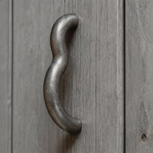 May include: A curved, silver-toned metal cabinet handle mounted on a gray wooden cabinet door. The handle has a textured, wavy design and is attached to the door with a single screw. The wood grain is visible.