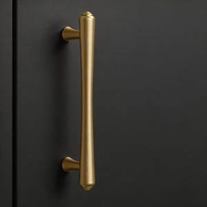 May include: A gold-colored cabinet pull with a vertical, elongated design. The pull has a cylindrical center that tapers at both ends, with a small, rounded base and top. The pull is mounted on a dark gray cabinet door.