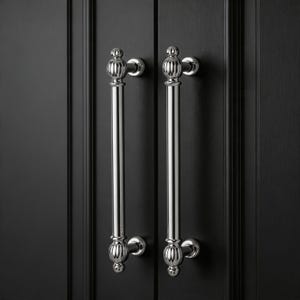 May include: Two polished chrome cabinet pulls with a vertical, elongated design. Each pull features decorative, rounded ends and a central, straight bar. The pulls are mounted on a black cabinet door.