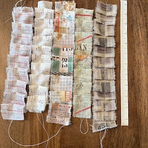 May include: A collection of fabric strips with frayed edges, featuring various patterns and text. The strips include musical notes, maps, and handwritten numbers. A ruler is placed next to the strips, with measurements in inches.
