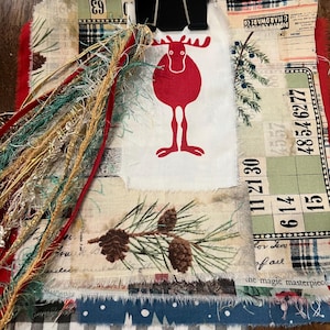 May include: A fabric collage featuring a central white patch with a red moose illustration, layered over various patterned textiles including pine cones, bingo cards, and plaid. A black metal binder clip holds the layers, adorned with decorative fringe.