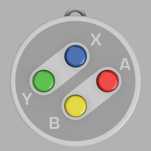 Könnte beinhalten: Ein grauer Anhänger mit dem Tastenlayout eines klassischen Videospiel-Controllers. Er hat vier farbige Knöpfe: grün, blau, rot und gelb, mit weißen Buchstaben. Der Anhänger hat oben eine Schlaufe zum Befestigen.
