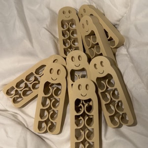 May include: A collection of beige-colored, smiling, decorative hangers. Each hanger has a rounded top with a smiling face and a series of circular cutouts down the body. The hangers are arranged on a white, textured surface.
