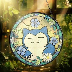 以下が含まれることがあります： 青いスノーラックスのキャラクターが目を閉じており、青い花、白いデイジー、緑の葉に囲まれた丸いステンドグラスの窓。窓は暗い縁取りがあり、チェーンから吊り下げられています。