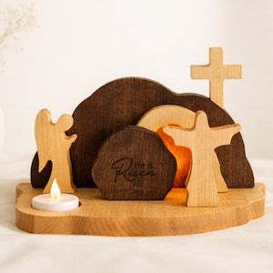 Può includere: Scena pasquale in legno con croce, angelo e figura con le braccia tese. La scena comprende una montagna marrone scuro, una croce marrone chiaro e una pietra con la scritta "He is Risen". Una piccola candela è accesa.