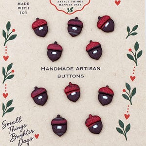 Könnte beinhalten: Zehn eichelförmige Knöpfe auf einer beigen Karte mit dem aufgedruckten Text MADE WITH JOY, ARTFUL THINGS HAPPIER DAYS, HANDMADE ARTISAN BUTTONS und Small Things Brighter Days. Jeder Knopf hat eine dunkelbraune Basis und eine glitzernde rote Kappe.
