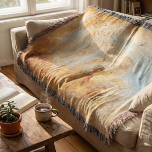 May include: A woven throw blanket draped over a beige sofa. The blanket features a landscape design with warm tones of brown, yellow, and blue, with a fringed edge. A small wooden table with a cup of tea and a potted plant is in the foreground.