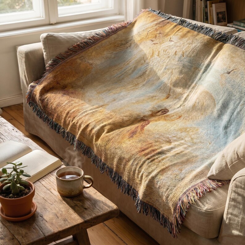 May include: A woven throw blanket draped over a beige sofa. The blanket features a landscape design with warm tones of brown, yellow, and blue, with a fringed edge. A small wooden table with a cup of tea and a potted plant is in the foreground.