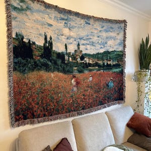 May include: A woven tapestry depicting a landscape scene with a field of red flowers, figures, and a village under a cloudy sky. The tapestry has a fringed border and is hung on a wall above a beige sofa.