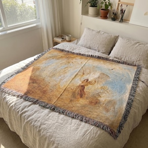May include: A rectangular woven throw blanket with a detailed painting of a figure in a field with birds and a blue sky. The blanket has a fringed edge and is primarily tan, brown, and blue.