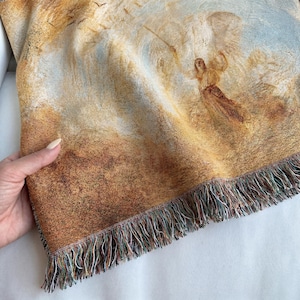 May include: Woven throw blanket featuring a classical painting of an angel in a landscape with a warm color palette of browns, tans, and blues. The blanket has a fringed edge.