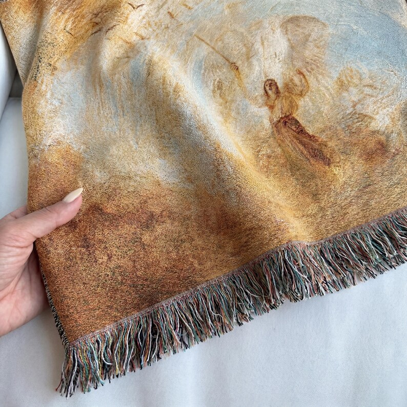 May include: Woven throw blanket featuring a classical painting of an angel in a landscape with a warm color palette of browns, tans, and blues. The blanket has a fringed edge.