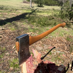 May include: A wood-handled axe with a steel blade is embedded in a piece of wood. The axe has a curved wooden handle and a silver-colored blade. The wood is a mix of pink and brown colors, with a rough texture. The background is a grassy area.