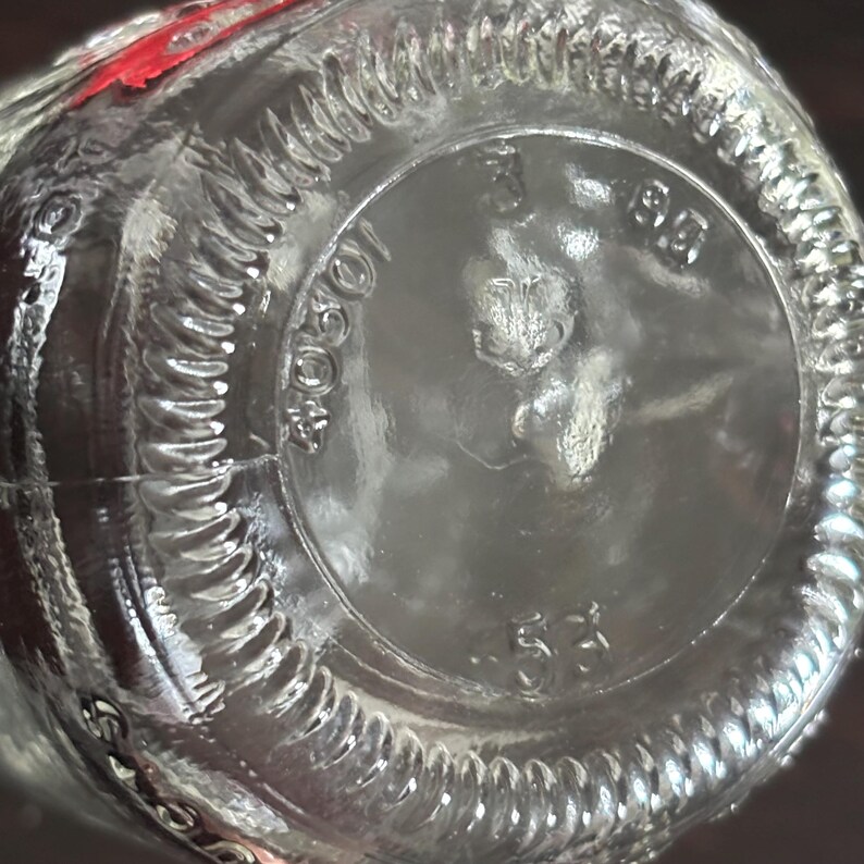 Close-up of the bottom of a clear glass mini mason jar. The base has a circular design with embossed text and numbers, including "1060401" and "53". The rim of the bottle has a textured, ridged pattern.