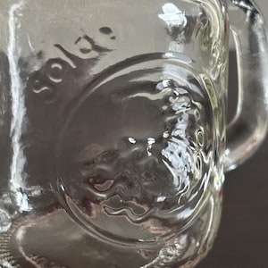 Clear glass mini mason jar spice shaker with a handle. The glsss has a circular embossed design and the word "golden" is pressed on the side. The glass is transparent, allowing light to pass through, and the handle is sturdy.