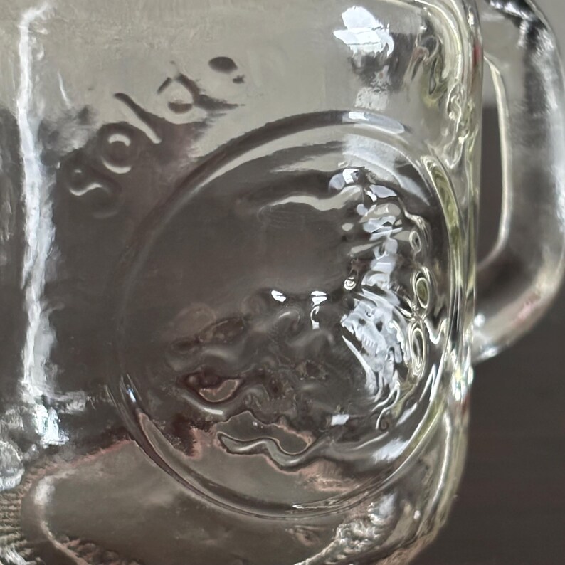 Clear glass mini mason jar spice shaker with a handle. The glsss has a circular embossed design and the word "golden" is pressed on the side. The glass is transparent, allowing light to pass through, and the handle is sturdy.