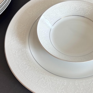 May include: A white porcelain dinnerware set featuring a detailed embossed floral pattern. The set includes a large plate and a smaller bowl, both with a delicate silver trim. The design is elegant and suitable for formal dining.