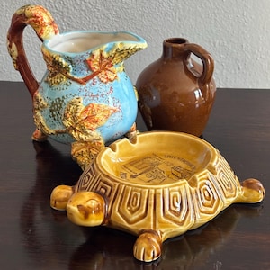 May include: A collection of vintage ceramic items. Includes a blue and brown pitcher with leaf details, a brown jug, and a turtle-shaped ashtray with a map design. The ashtray is yellow and brown.