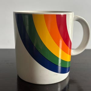 May include: White ceramic mug with a curved rainbow design in red, orange, yellow, green, and blue. The mug has a curved handle and is suitable for hot or cold beverages.