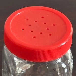 Close-up of a clear glass jar with a bright red plastic lid. The lid has multiple small holes and a textured, ridged edge. The jar appears to be empty.
