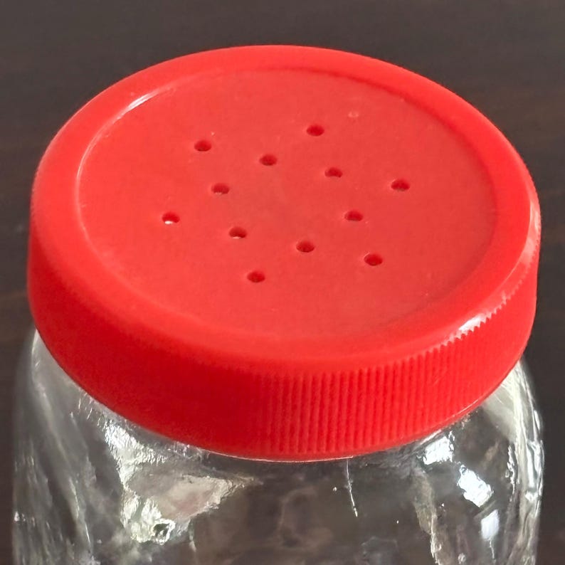 Close-up of a clear glass jar with a bright red plastic lid. The lid has multiple small holes and a textured, ridged edge. The jar appears to be empty.
