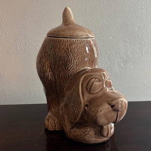 May include: A brown ceramic dog-shaped cookie jar with a lid. The jar is a light brown color with a textured surface, and the dog's face has a friendly expression. The jar is sitting on a dark brown surface.