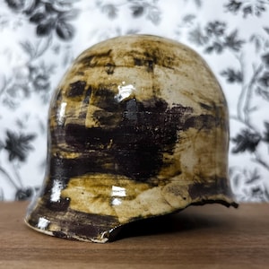 May include: A ceramic bell-shaped object with a glossy finish. The bell has a beige base color with dark brown and olive green brushstrokes. The object sits on a wooden surface against a floral patterned background.