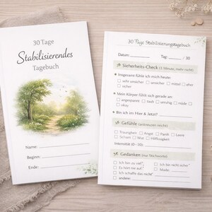 May include: A white journal with the German text "30 Tage Stabilisierendes Tagebuch" and a watercolor illustration of a path through a forest. A pen and an open page with checklist prompts are also visible.
