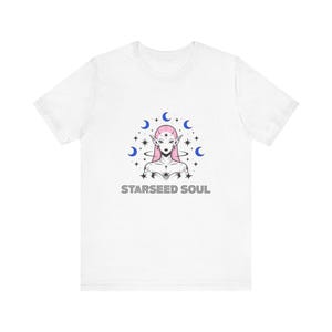 May include: White t-shirt featuring a graphic of an ethereal figure with pink hair and pointed ears, surrounded by blue crescent moons and stars. The text "STARSEED SOUL" is printed below the graphic in a sans-serif font.