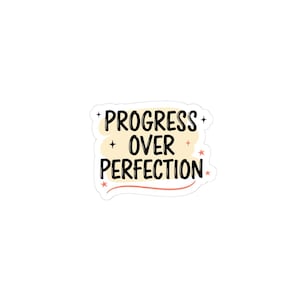 May include: A white sticker featuring the words "PROGRESS OVER PERFECTION" in black, handwritten font. The text is set against a pale yellow backdrop, with small star embellishments.