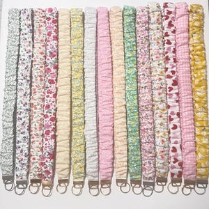 Puede incluir: Una colección de cordones de tela floral en varios colores y patrones. Cada cordón presenta un anillo y un cierre de metal. Los diseños incluyen pequeñas flores, corazones y patrones de cuadros en tonos de rosa, amarillo y verde.