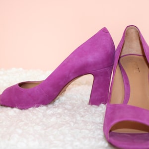 May include: A pair of bright purple suede peep-toe heels. The shoes have a chunky heel and a rounded toe. The interior of the shoe is a light tan color. The shoes are displayed on a white, fluffy surface against a light pink background.