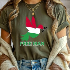 May include: Olive green t-shirt featuring a dove design with the colors of the Iranian flag. The dove holds an olive branch and the text "FREE IRAN" is printed below the dove. The shirt is made of a soft material.