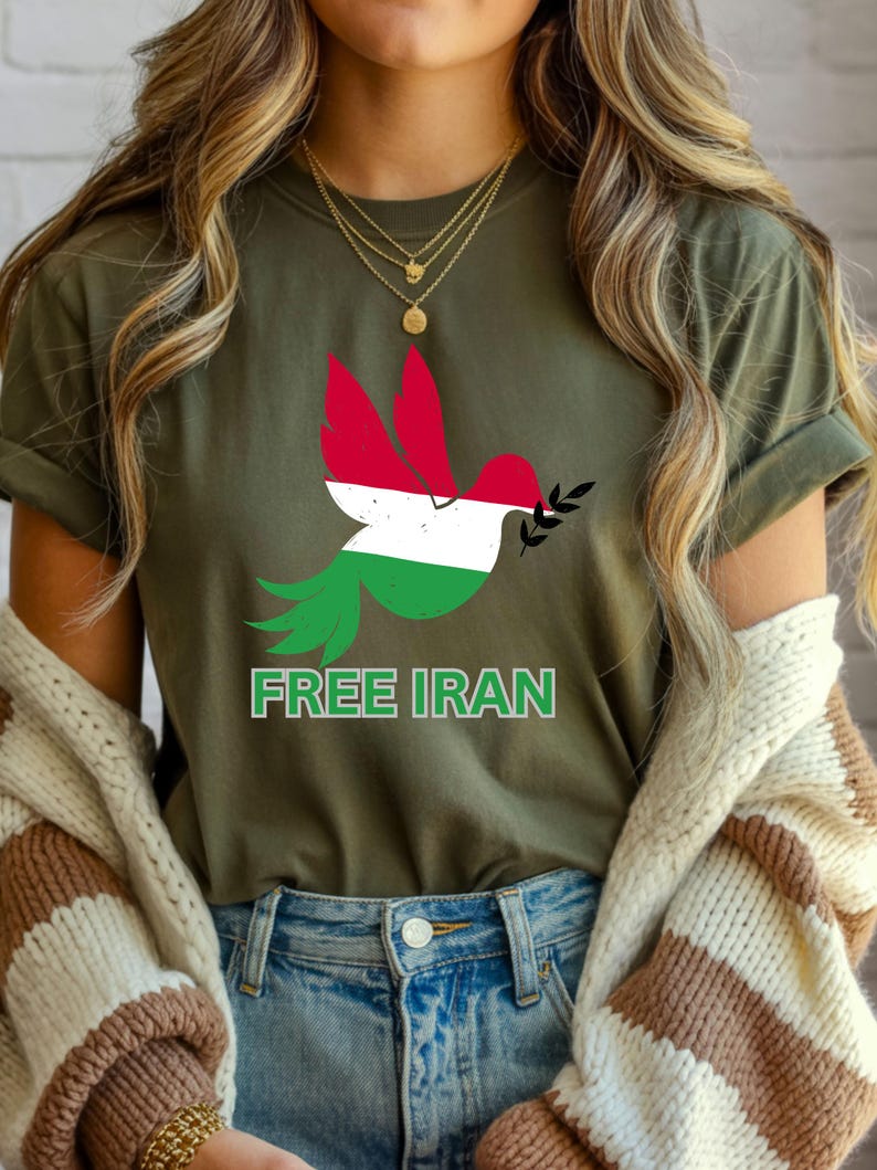May include: Olive green t-shirt featuring a dove design with the colors of the Iranian flag. The dove holds an olive branch and the text "FREE IRAN" is printed below the dove. The shirt is made of a soft material.