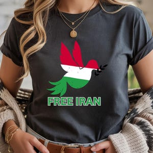 May include: Dark gray t-shirt featuring a dove with wings in the colors of the Iranian flag, holding an olive branch. The text "FREE IRAN" is printed in green below the dove. The shirt has rolled-up sleeves.