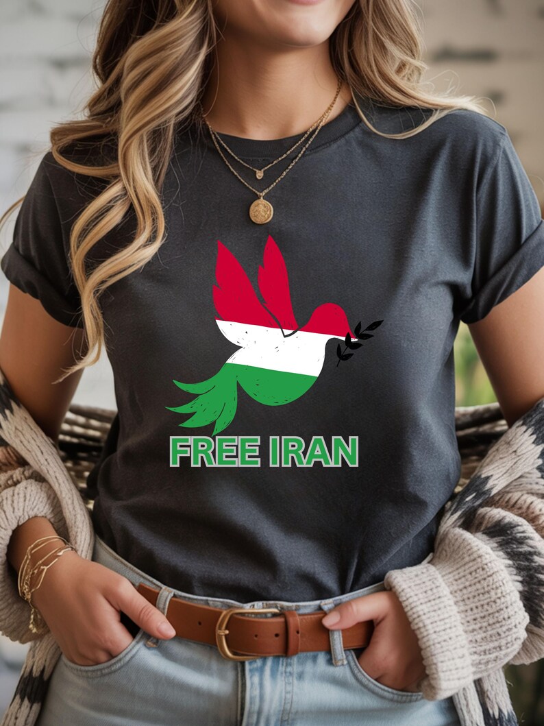 May include: Dark gray t-shirt featuring a dove with wings in the colors of the Iranian flag, holding an olive branch. The text "FREE IRAN" is printed in green below the dove. The shirt has rolled-up sleeves.