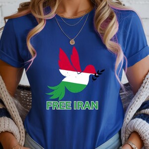 May include: A blue t-shirt featuring a dove with wings in the colors of the Iranian flag, holding an olive branch. The text "FREE IRAN" is printed in green below the dove. The shirt is a crew neck.