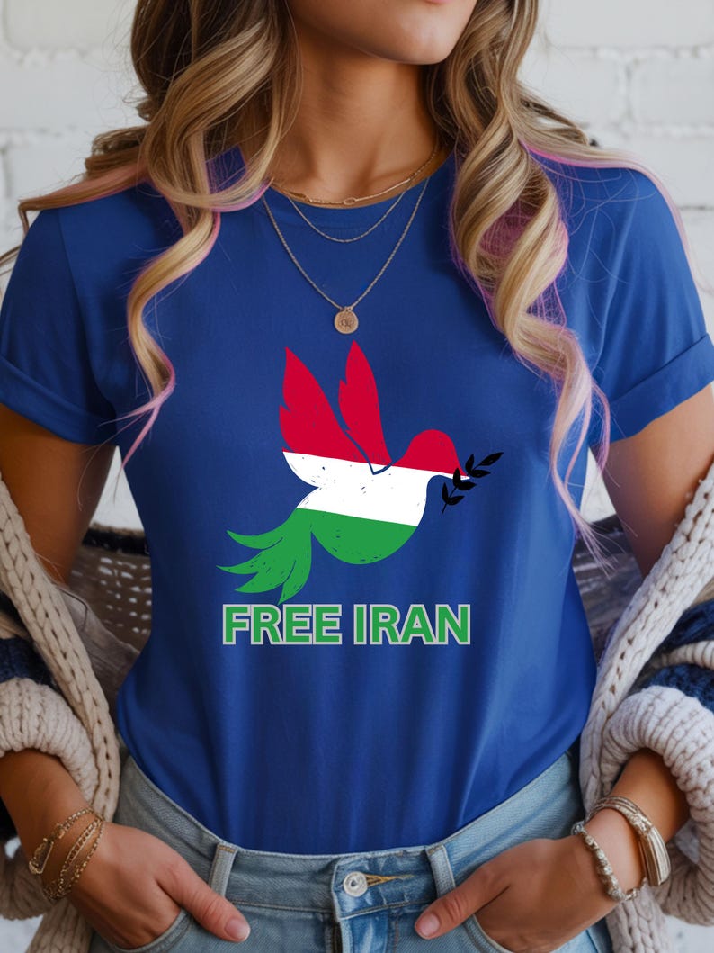 May include: A blue t-shirt featuring a dove with wings in the colors of the Iranian flag, holding an olive branch. The text "FREE IRAN" is printed in green below the dove. The shirt is a crew neck.