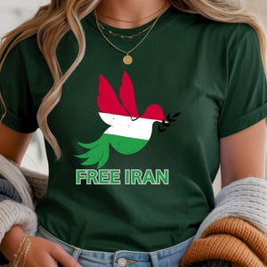 May include: Dark green t-shirt featuring a dove with red, white, and green colors, holding an olive branch. The text "FREE IRAN" is printed in green below the dove. The shirt is a crew neck.