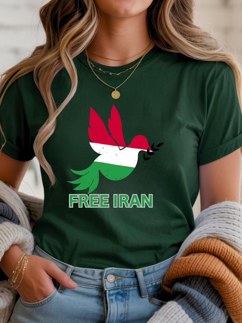 May include: Dark green t-shirt featuring a dove with red, white, and green colors, holding an olive branch. The text "FREE IRAN" is printed in green below the dove. The shirt is a crew neck.